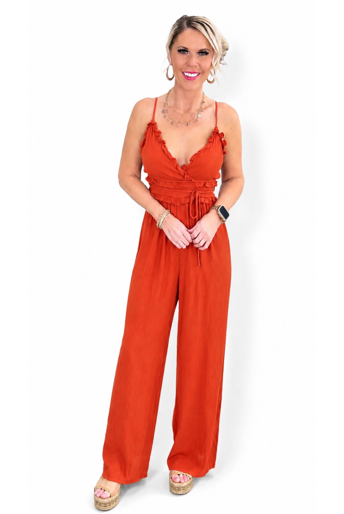 A woman modeling a bright rust-orange jumpsuit with thin straps, ruffled trim, a tie waist, and wide legs.