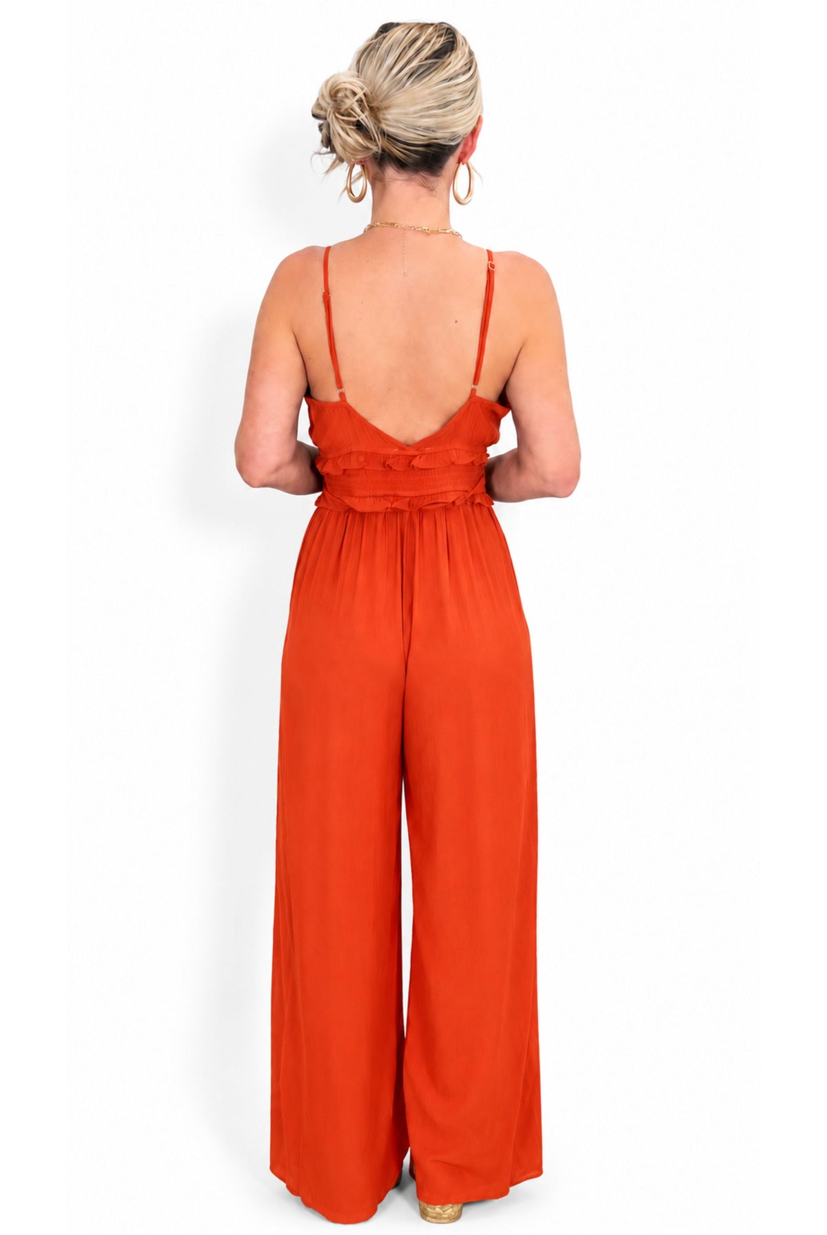 A woman viewed from the back wearing a bright orange wide-leg jumpsuit with thin straps and a ruffled back detail.