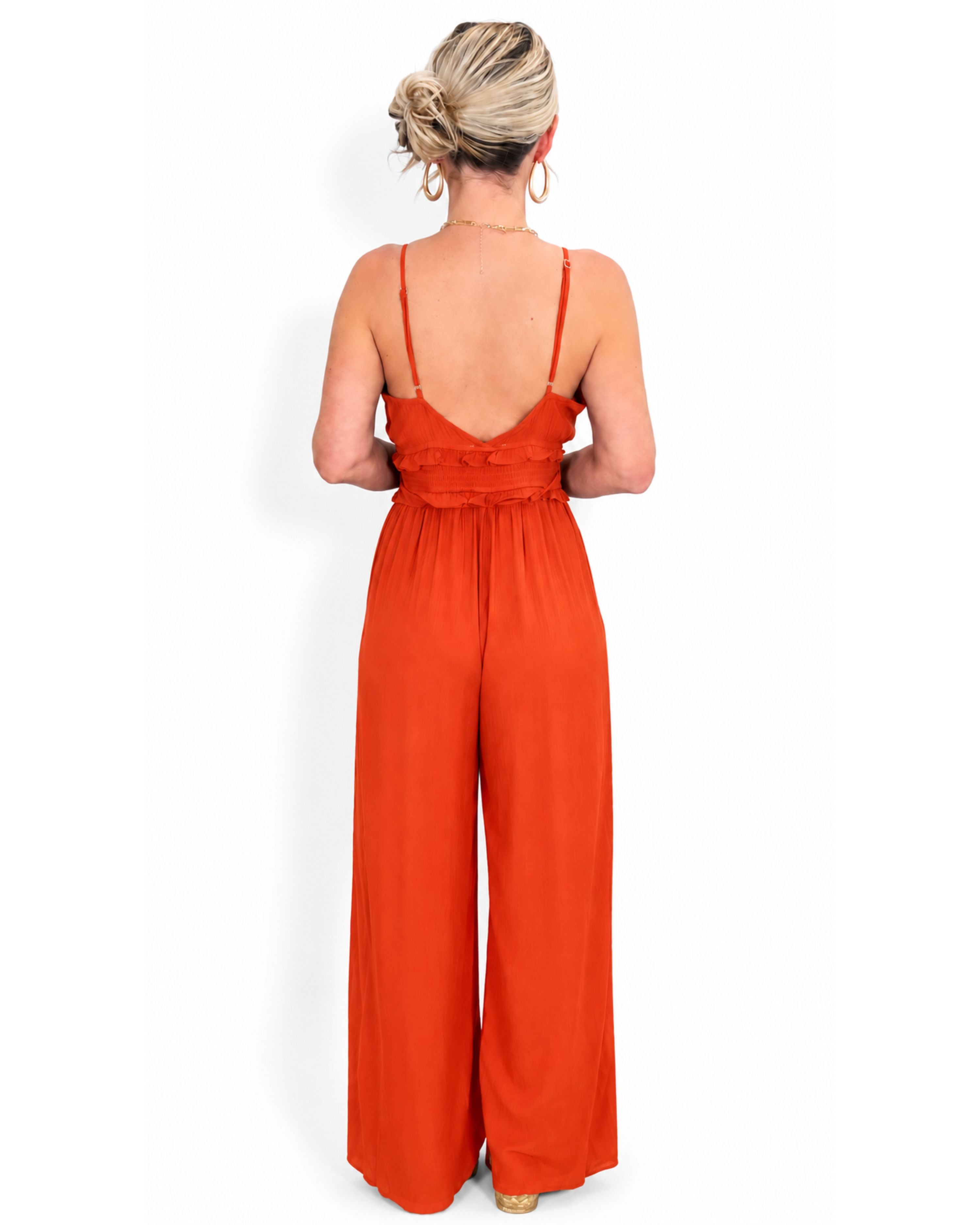 A woman viewed from the back wearing a bright orange wide-leg jumpsuit with thin straps and a ruffled back detail.