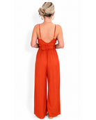 A woman viewed from the back wearing a bright orange wide-leg jumpsuit with thin straps and a ruffled back detail.