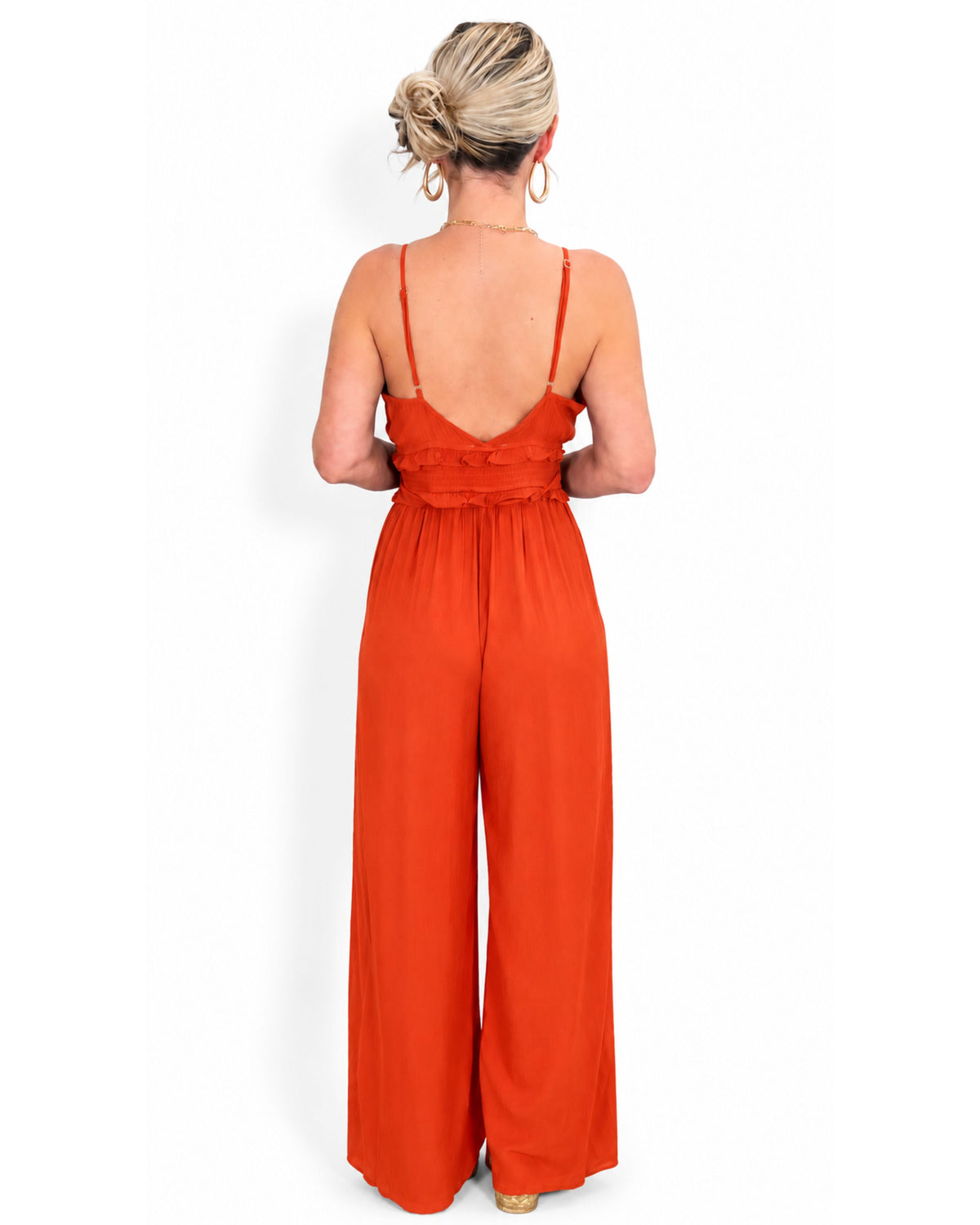 A woman viewed from the back wearing a bright orange wide-leg jumpsuit with thin straps and a ruffled back detail.